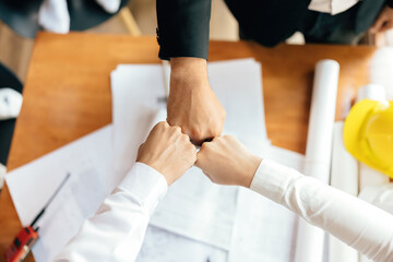 An image of 3 people joining hands of the architecture team. Represents unity and energy in work. There is a blueprint of an engineering design project on a desk in an office.