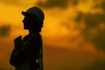 Half body silhouette portrait of smiling female engineer golden sky at sunset stand holding tablet Wear a helmet and a vest. is an inspection manager of an industrial factory about energy and safety