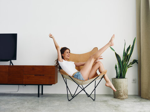 Woman lying in a leather armchair raised arms and legs cheerful happiness and a smile, relaxing at home stylish modern interior with white walls, copy space.