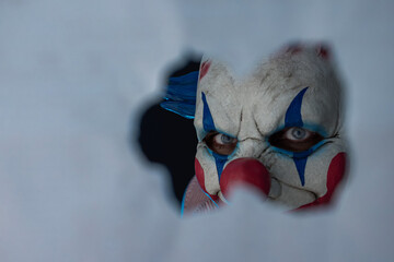 Creepy clown face peering through a hole in the partition. The face of a man wearing a creepy clown...
