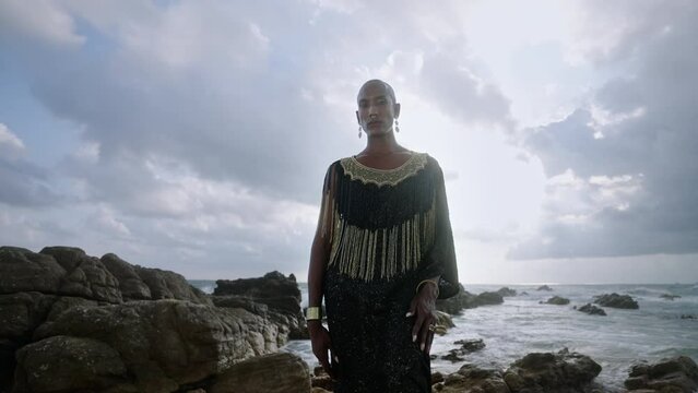 Gorgeous gay black man in luxury gown poses on scenic ocean beach slowmo. Non-binary ethnic fashion model in long posh dress and accessories looks at camera, dances with hands in slow motion