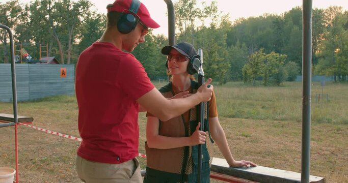 handsome caucasian man in headset is confidently teaching woman to use rifle, weapon on outdoor range, Handsome male and pleasant lady in protective clothes equipment outfit learning to shoot, talking