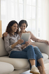 Obraz premium Cheerful adult child woman showing online content on tablet to cheerful senior mom, talking on video call, enjoying Internet technology, communication, relaxing together at home
