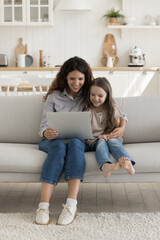 Happy excited mom and cheerful kid girl using learning app on laptop for preschooler education, enjoying domestic internet technology, communication, sitting on sofa with gadget