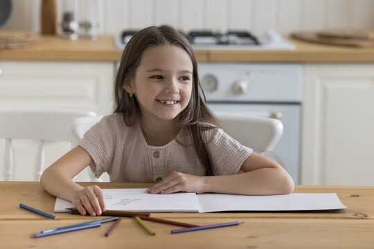 Happy joyful little kid drawing in coloring pencils in paper album at home, sitting at kitchen table, looking away, smiling, laughing. Kid engaged in learning game, developing creative artistic skills