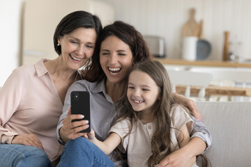 Joyful kid girl, mom, grandma relaxing on home sofa with smartphone, using app, media service for...