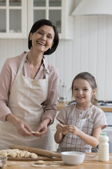 Cheerful pretty grandmother and sweet kid girl in aprons baking dessert t kitchen table together, shaping dumplings, pies, cookies over floury table, looking at camera, smiling, laughing