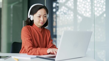 Asian woman in wireless headphones IT professional programmer working typing coding on laptop while sitting at workplace in modern office. Busy businesswoman developer analyzes financial digital data - Powered by Adobe