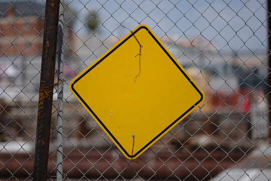 A blank, yellow diamond-shaped signs functions as a warning sign. It is tied onto a chainlink fence at a construction site for a major infrastructure project.