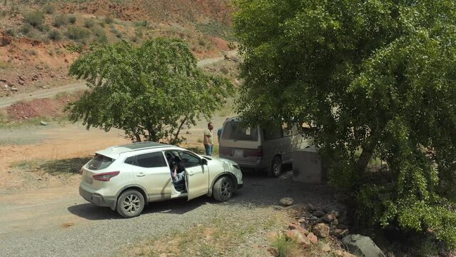Travelers stopped by cars next to river and trees in picturesque place in mountains and communicate cheerfully communicate, circular shooting from above with drone. Concept of travel and adventure.
