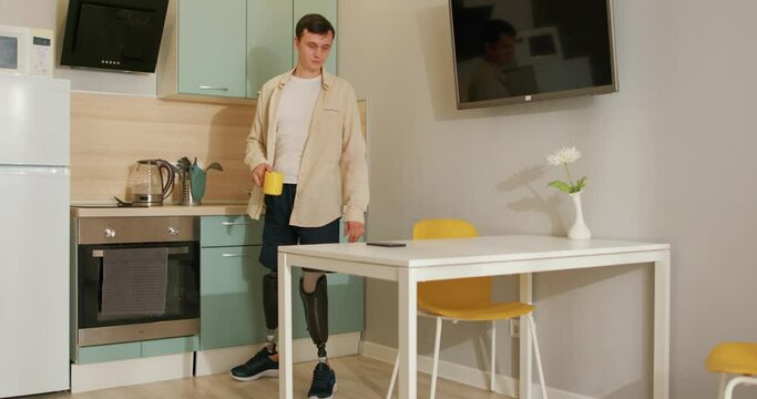 handsome disabled man with bionic smart legs prosthesis sit in kitchen, watching movie on smartphone and drinking coffee or tea, alone, indoors. physical disability, people concept