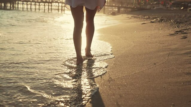 Legs of woman tourist barefooted feet walking on sandy ocean beach leaving footprints at sunset, rear view. Female wearing airy white tunic. Enjoying resting relaxing on vacation. Travel, tourism.