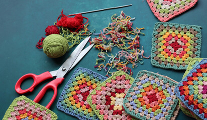 Fototapeta premium Red scissors, cotton yarn balls, crochet hook and granny squares on a blue desk. Messy working place. Crochet geometric shape. 