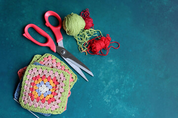 Red scissors, cotton yarn balls, crochet hook and granny squares on a blue desk. Messy working...