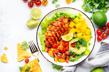 Yummy salad with grilled chicken fillet with mango, spicy salsa, tomatoes, cilantro, red onion and lettuce in tex-mex style, white table background, top view
