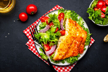 Fried white fish fillet with vegetable salad from lettuce, cherry tomatoes and red onion, black table background, top view - obrazy, fototapety, plakaty