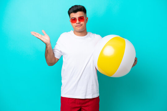 Young caucasian man holding a beach ball isolated on blue background having doubts while raising hands