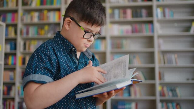 Side portrait of kid boy with Down syndrome, leafing through the pages while reading book in library