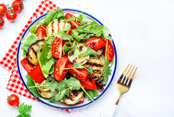 Grilled vegetables salad with paprika, zucchini, eggplant, tomatoes and spinach on plate, white table background, top view