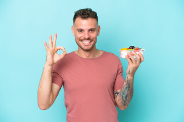 Young caucasian man holding a bowl of fruit isolated on blue background showing ok sign with fingers