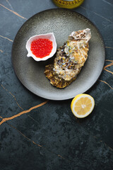 Olive-colored plate with fresh unopened oyster and red caviar, flat lay on a dark-olive marble background, vertical shot with copy space