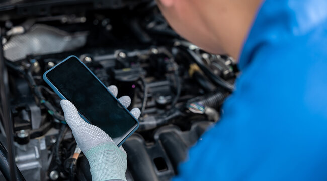Mechanic taking control of car repair with smartphone at workshop. Close-up shot of male technician examining engine with wrench. Horizontal photo with garage background.