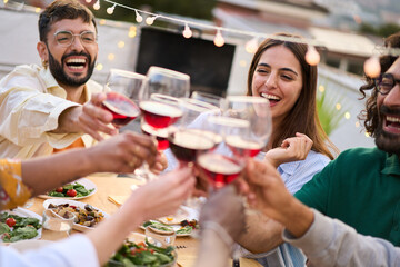 Gathering of cheerful young friends toasting with red wine and celebrating party on terrace of...