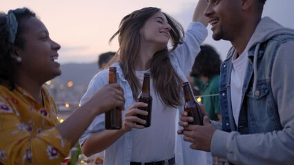Happy young multiracial group dancing friends at rooftop BBQ party. Excited people jumping have fun drinking beer at barbecue in evening. Students celebrating summer vacation. - Powered by Adobe