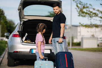 A little girl carries her suitcase to the car. Dad helps his daughter put the luggage in the trunk