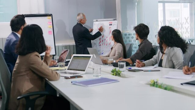 An Indian Asian old male corporate businessman delivering presentation in an interactive brainstorming office session using white charts graphs with team of employees at a conference boardroom meeting
