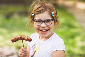 Little preschool girl eating grilled sausage. Happy child on barbecue or picknick. Healthy food,...