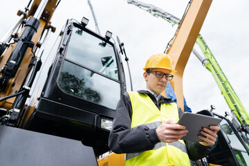 Engineer in a helmet with a digital tablet next to construction excavator