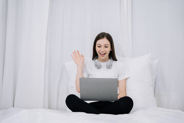 Happy woman having fun doing video call using laptop in pajama relaxing lying on a bed.