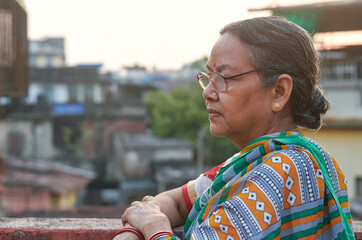 Photo of a simple looking aged Bengali woman (Indian ethnicity) with saddened / depressed look (feeling of loneliness) in her face. Photo taken in a building rooftop, in Kolkata, West Bengal. - obrazy, fototapety, plakaty