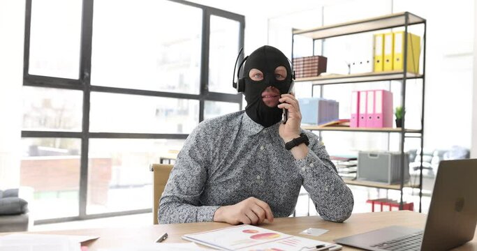 Fake manager in mask of robber talks to client sitting at workplace in company premise. Male scammer in headphones communicates cheating customer via call