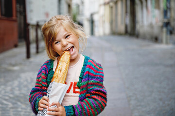 Adorable little preschool girl with fresh French baguette on the street side of the city. Happy...