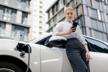 Closeup progressive businesswoman talking on smartphone, leaning electric car at charging station...