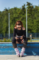 Inline skater female sitting on a ledge in a skatepark and playing online video game on a smartphone with a gamepad. Cool young roller blader person gaming online with a modern gadget