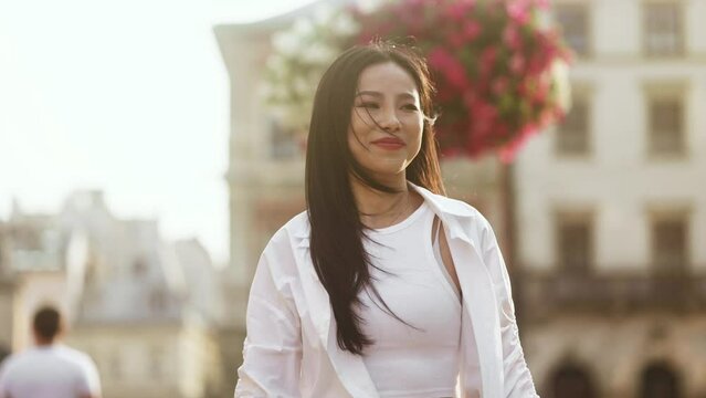 Charming young asian woman walking down the street with flying dark hair and looking around outdoors Happy relaxed lady walking on the city centre enjoying beautiful day