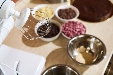 Selective focus on a whisk of food processor on the kitchen countertop with blurred organic dark, caramel, white and ruby pink chocolate chips. Ingredients for making glaze, sweet desserts, brownies