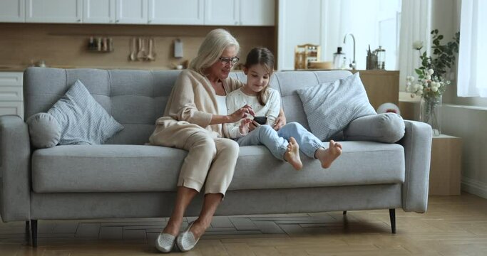 Mature 60s granny and little 6s granddaughter using cellphone at home, laugh, look at gadget screen, watch videos, spend leisure on internet, play cool modern videogames, enjoy on-line vlog content