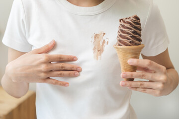 Cloth stain, disappointment asian young woman, girl eating melting ice cream in waffle cone on hot weather, hand show making chocolate cream drop on white t-shirt, spot dirty or smudge on clothes. - Powered by Adobe