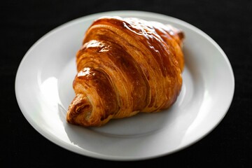 Closeup shot of a single croissant on a white plate in the black background.