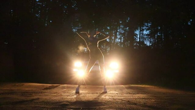 A young slender girl dances a modern dance against the background of car headlights in a night forest