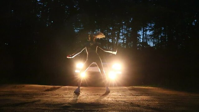 A young slender girl dances a modern dance against the background of car headlights in a night forest