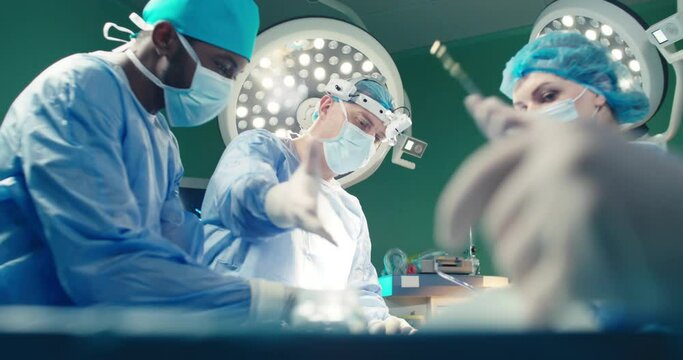 Diverse team of professional surgeons performing invasive surgery on patient in hospital operating room. African American assistant hands out instruments to surgeon. Ethnic diversity.