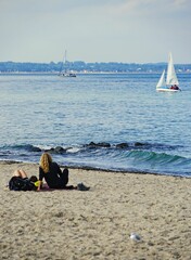 Vertical back view of people resting on sandy Travemuende beach with sailboats in the background © Stephan Hickisch/Wirestock Creators