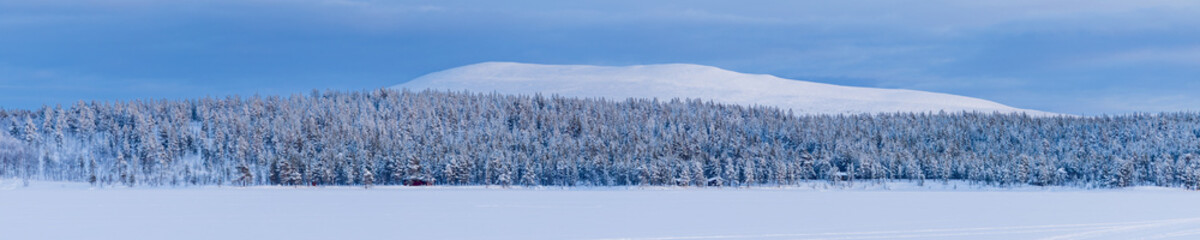 Panoramic photo of the snow capped Pyhakero mountain in Lapland, Finland with a pine forest and frozen lake in the foreground