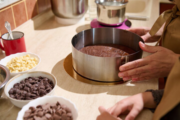 Close-up baker confectioner putting a chocolate biscuit inside a detachable metal cake mold, standing at kitchen counter with delicious food ingredients. Bakery. Confectionery. Culinary concept