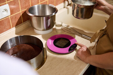 Close-up of pastry chef, confectioner, standing by marble kitchen countertop, using kitchen scale weighing food ingredients while making homemade sweet dessert. Culinary. Bakery. Confectionery